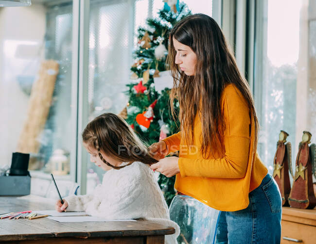 Side view of female adolescent braiding hair of sibling drawing on paper sheet with pencil near Christmas tree at home — Stock Photo
