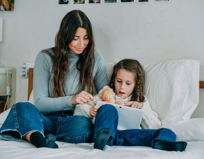 Full body smiling young woman in casual wear showing mobile phone screen to cute positive daughter while sitting together on comfy bed — Stock Photo