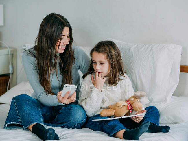 Ganzkörperlächelnde junge Frau in Freizeitkleidung, die der netten positiven Tochter ihren Handy-Bildschirm zeigt, während sie zusammen auf einem bequemen Bett sitzt — Stockfoto