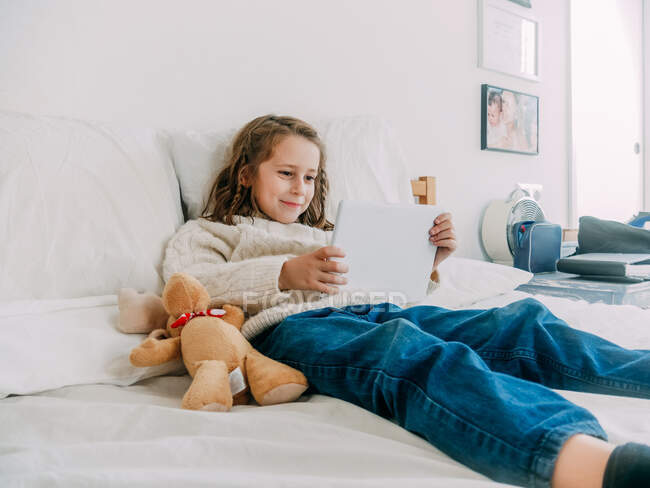 Lächelnd fröhliches kleines Mädchen in lässiger Kleidung auf bequemem Bett liegend und Video auf modernem Tablet im hellen Schlafzimmer anschauend — Stockfoto