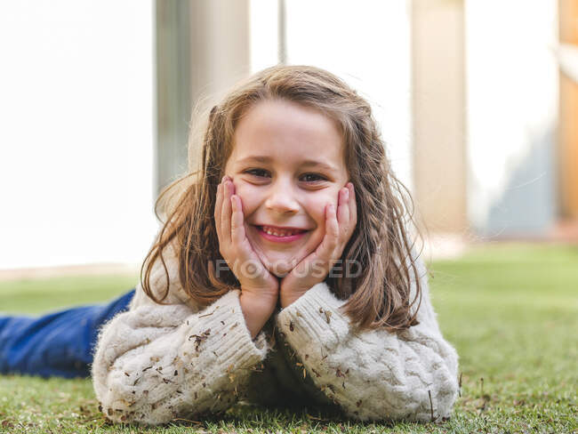 Очаровательная веселая маленькая девочка в уютном белом свитере лежит на травянистом лугу и смотрит в камеру с улыбкой — стоковое фото