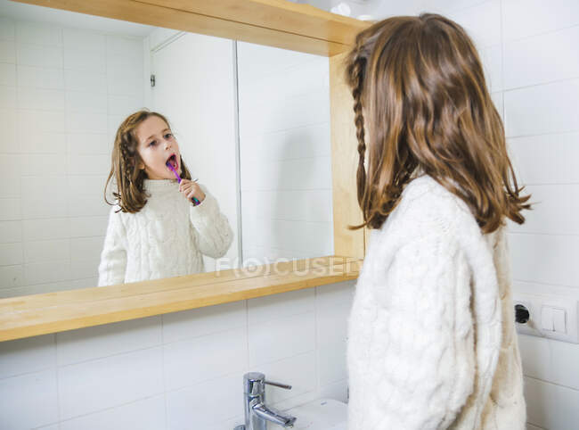 Vista lateral niña feliz vistiendo suéter blanco acogedor mirando en el espejo con sonrisa dentada mientras está de pie con cepillo de dientes en el baño moderno - foto de stock
