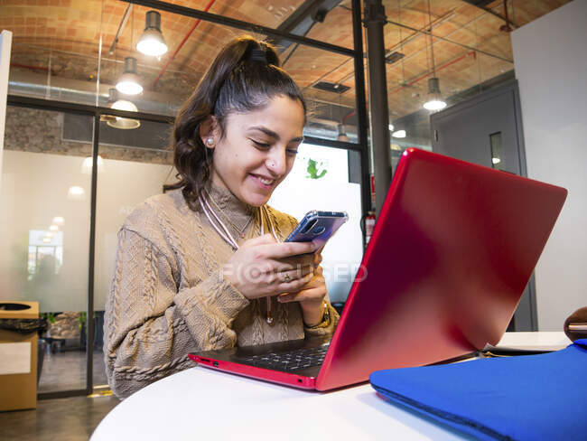 Low angle of smiling female student text messaging on cellphone while using laptop for studies in university — Stock Photo