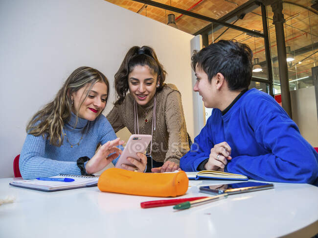 Group of smiling female classmates gathering around table with stationery and notebooks for searching information in cellphone for homework task — Stock Photo