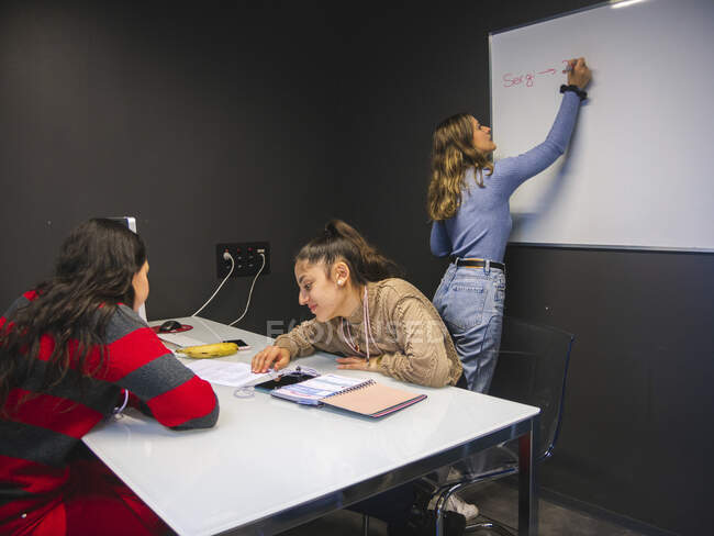 Mujer aprendiz escribiendo con marcador en pizarra blanca mientras sus compañeras de clase trabajan en el proyecto sentadas en la mesa en el aula universitaria - foto de stock