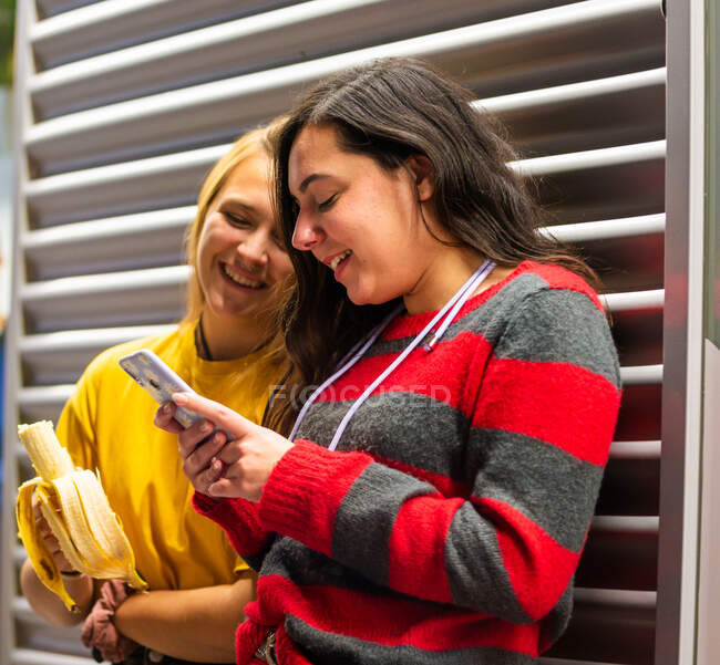 Fröhliche Studenten, die während ihres Studiums eine Pause einlegen und Bananen essen, während sie auf dem Handy in den sozialen Medien surfen — Stockfoto
