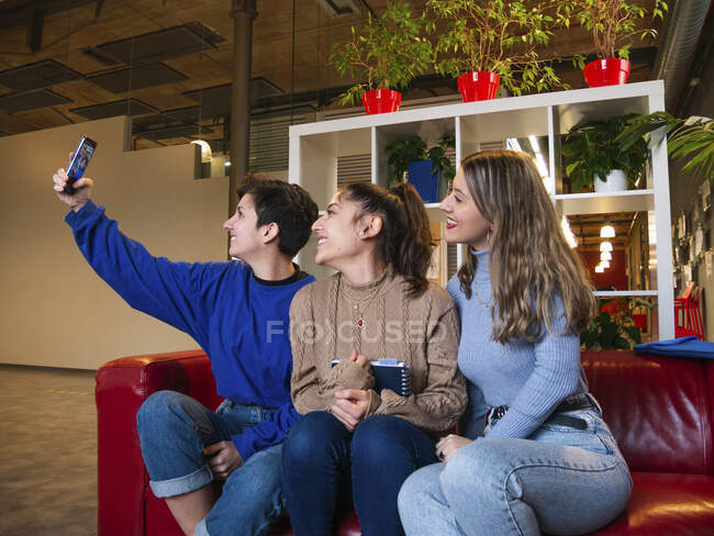Улыбающиеся студентки сидят на диване с ноутбуком и делают автопортрет на мобильном телефоне во время перерыва — стоковое фото