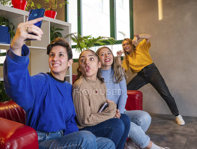 Estudiantes sonrientes sentadas en un sofá con portátil y tomando autorretrato en el teléfono móvil durante el descanso - foto de stock