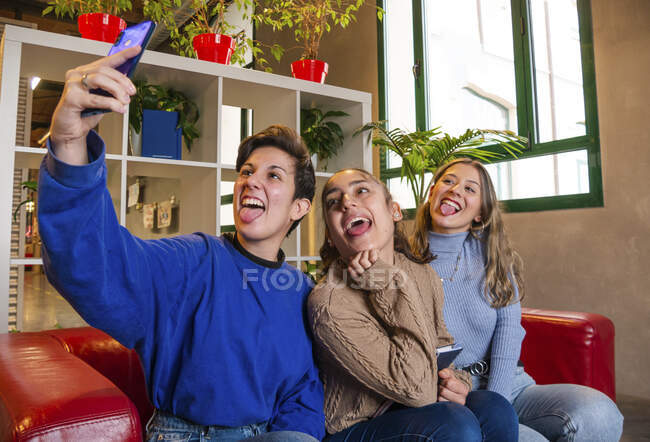 Studenti donne sorridenti sedute sul divano con taccuino e che scattano autoritratti sul cellulare durante la pausa — Foto stock