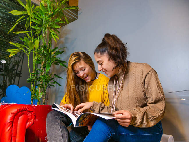 Positive Freundinnen sitzen eng beieinander und lesen Informationen im Album, während sie ihre Freizeit miteinander verbringen — Stockfoto