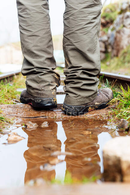 Visão traseira pessoa anônima em calças cinza em pé na estrada de ferro em terreno rochoso e refletindo em poça no campo — Fotografia de Stock