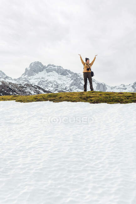 Cuerpo entero alegre hembra en ropa de abrigo levantando los brazos en la emoción mientras está de pie en el vasto valle montañoso cubierto de nieve y rodeado de montañas rocosas severas - foto de stock