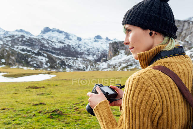 Vista lateral enfocada joven fotógrafa en suéter cálido tomando fotos en cámara fotográfica profesional de majestuosas montañas ásperas en tierras altas cubiertas de hierba en el día claro de otoño - foto de stock