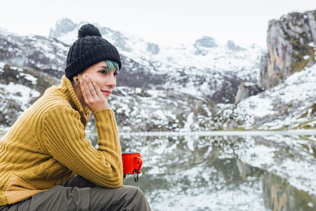 Vista lateral tranquila joven hembra en suéter cálido y sombrero beber bebida caliente mientras está sentado en piedra afilada en la orilla del lago frío rodeado de montañas nevadas severas ásperas - foto de stock
