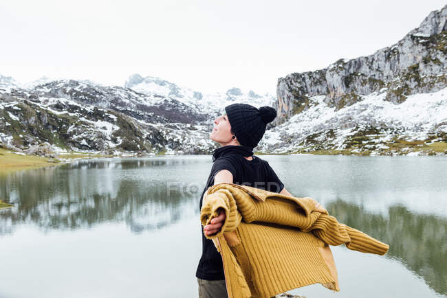 Back view attractive joyful female in warm sweater and hat outstretching arms in excitement and looking away while standing on cold rippling lake shore surrounded by rocky snowy mountains — Stock Photo