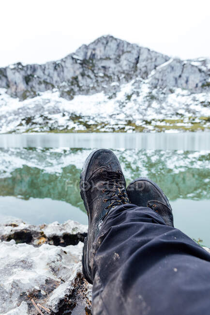 Crop anonymous traveler sitting with legs crossed on rough rocky terrain near cold lake against snowy majestic mountain — Stock Photo