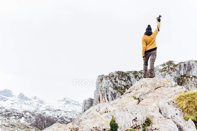 Back view full body photographer in warm clothes raising arm with photo camera and standing on stiff rough rock in snowy highlands in Asturias — Stock Photo