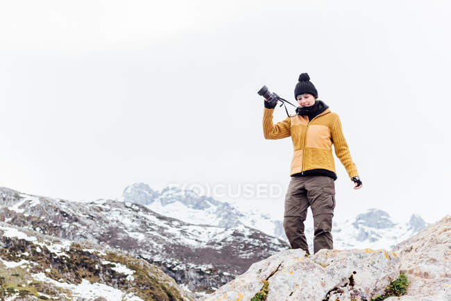 Fotógrafa de cuerpo entero en ropa de abrigo levantando el brazo con cámara fotográfica y de pie sobre roca rugosa rígida en tierras altas nevadas de Asturias - foto de stock