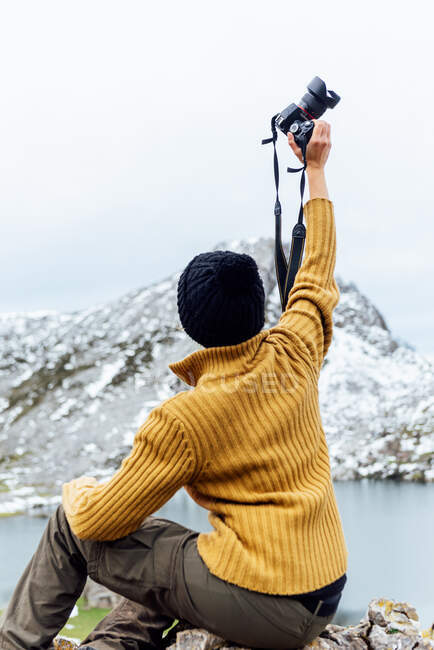 Фотограф на всьому тілі в теплому одязі з фотокамерою, сидячи на жорсткій грубій скелі в снігових високогір 