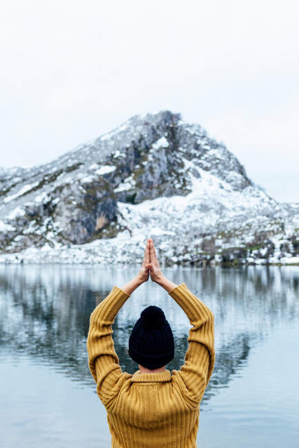 Back view unrecognizable female in warm clothes and hat meditating on Lotus Pose with Namaste hands on lakeside against severe snowy mountains in Asturias — Stock Photo