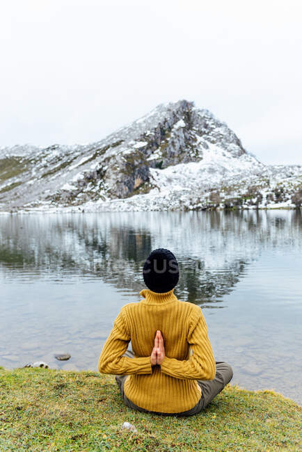 Вид сзади неузнаваемая женщина в теплой одежде и шляпе медитирует на Lotus Pose с Namaste руки позади на берегу озера против тяжелых снежных гор в Астурии — стоковое фото