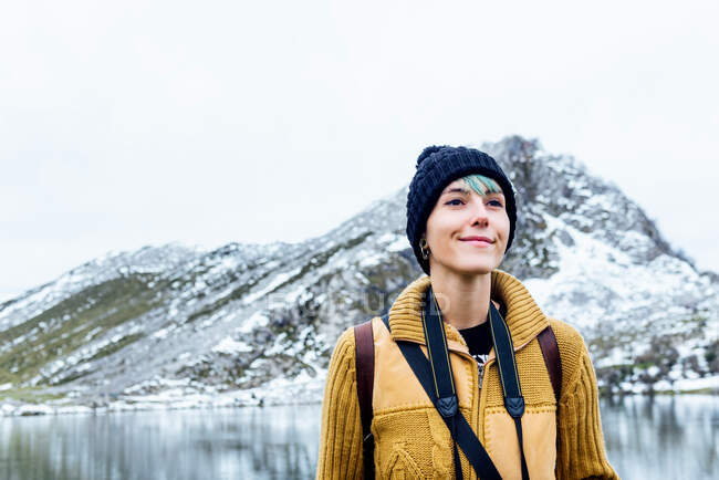 Jovem conteúdo viajante feminino em roupas quentes olhando para longe contra monte nevado refletindo no lago sob o céu branco na Espanha — Fotografia de Stock