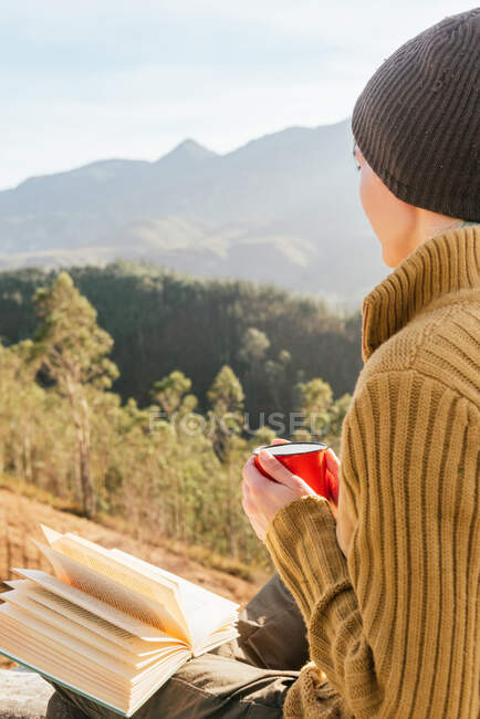 Боковой вид женщины, сидящей за чашкой горячего напитка и читающей интересную книгу на фоне живописного горного пейзажа в солнечный день — стоковое фото