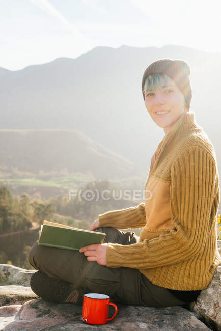 Вид збоку на зміст жінка мандрівник сидить з чашкою гарячого напою і читає цікаву книгу на тлі ефектного гірського пейзажу в сонячний день — стокове фото
