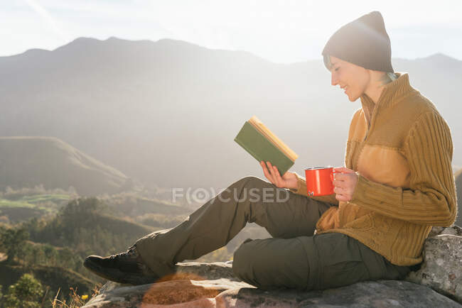 Vista lateral del contenido viajero femenino sentado con una taza de bebida caliente y leyendo un interesante libro sobre el fondo del espectacular paisaje montañoso en un día soleado - foto de stock