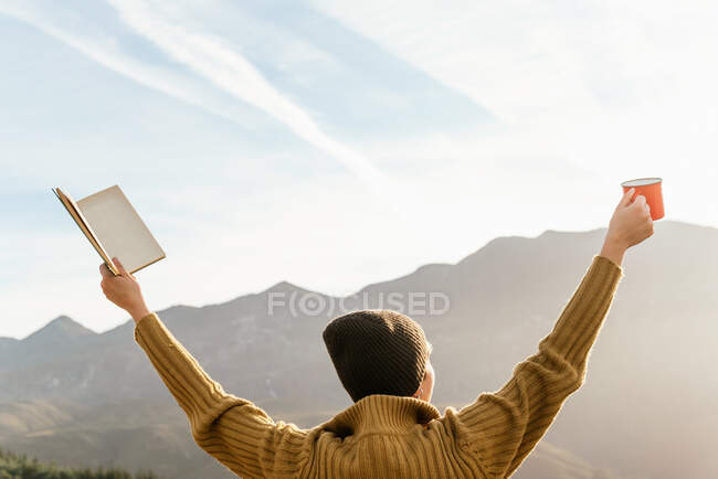 Обратный вид неузнаваемой женщины-исследователя, стоящей с книгой и чашкой горячего напитка в поднятых руках и наслаждающейся свободой в горах в солнечный день — стоковое фото