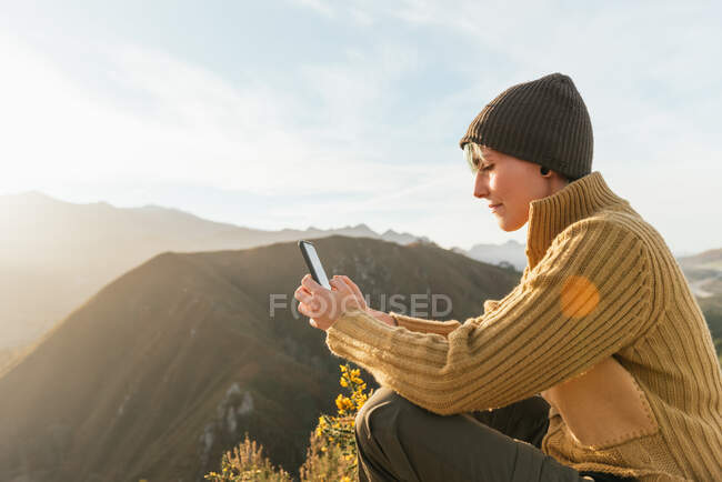 Vista laterale di messaggistica esploratore femminile positivo sul telefono cellulare mentre seduto sullo sfondo della catena montuosa nella giornata di sole — Foto stock