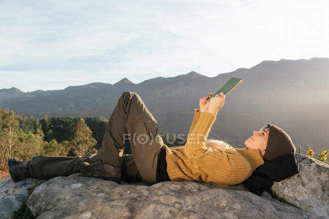Вид збоку на зміст жінка мандрівник лежить з чашкою гарячого напою і читає цікаву книгу на тлі ефектного гірського пейзажу в сонячний день — стокове фото