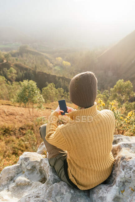 Back view of female explorer messaging on mobile phone while sitting on background of mountain range on sunny day — Stock Photo