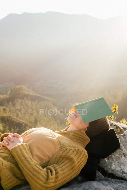 Side view of peaceful female traveler lying on rocks and covering face with book while sleeping in mountains on sunny day — Stock Photo
