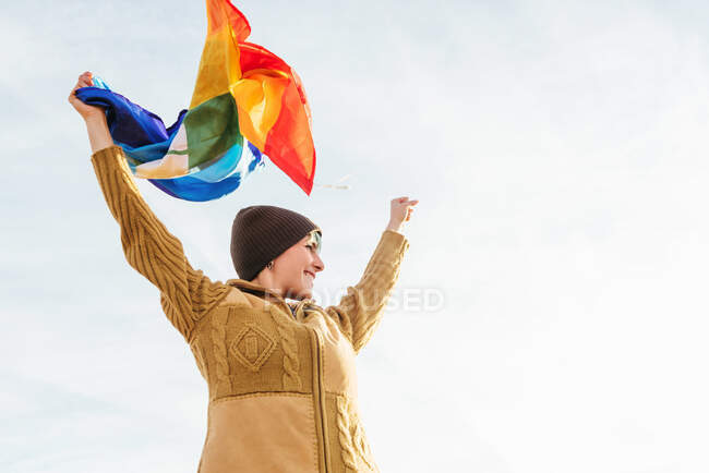 Desde abajo de excursionista de pie con arco iris bandera LGBT con inscripción Paz y disfrutar de la libertad con los brazos extendidos en las montañas - foto de stock