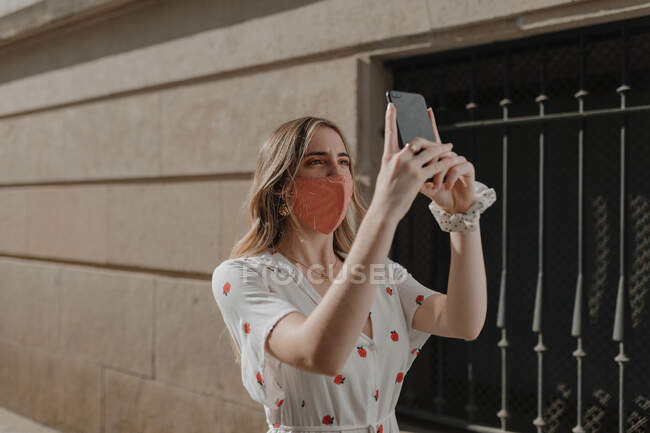 Jovem fêmea atenta anônima em máscara e desgaste ornamental tomando auto-retrato no celular na cidade durante a pandemia de COVID 19 — Fotografia de Stock