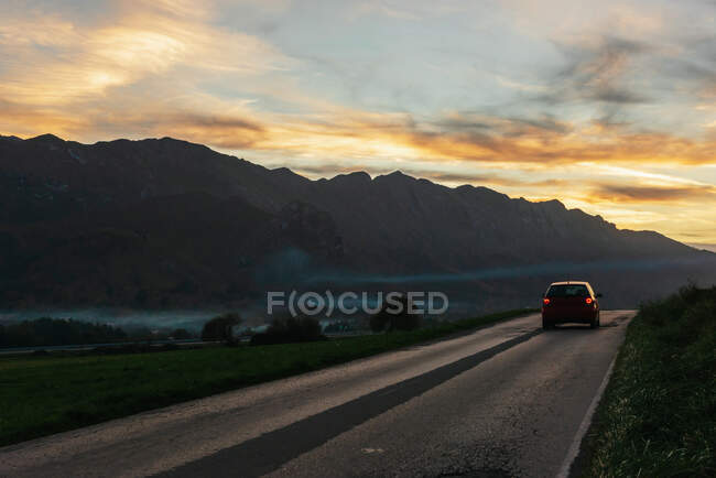 Automobile cavalcando lungo la strada asfaltata sullo sfondo della catena montuosa sotto il cielo al tramonto in serata — Foto stock