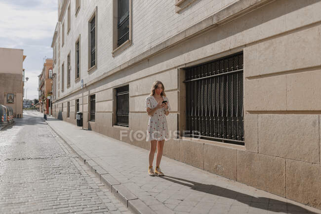 Unrecognizable female tourist in dress and mask text messaging on cellphone on pavement near stone building in Seville — Stock Photo