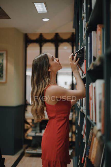 Vista laterale di giovane donna attenta in sundress rosso con capelli ondulati che sceglie il libro di testo dalla libreria con collezione di letteratura in libreria — Foto stock
