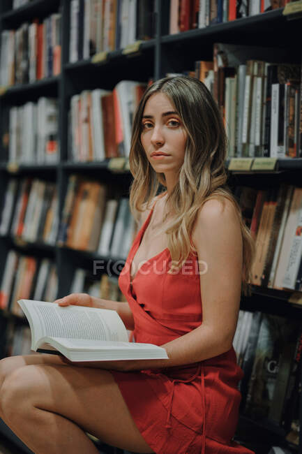 Vista laterale della giovane donna in prendisole rosso con libro di testo aperto seduta in libreria e guardando la macchina fotografica — Foto stock