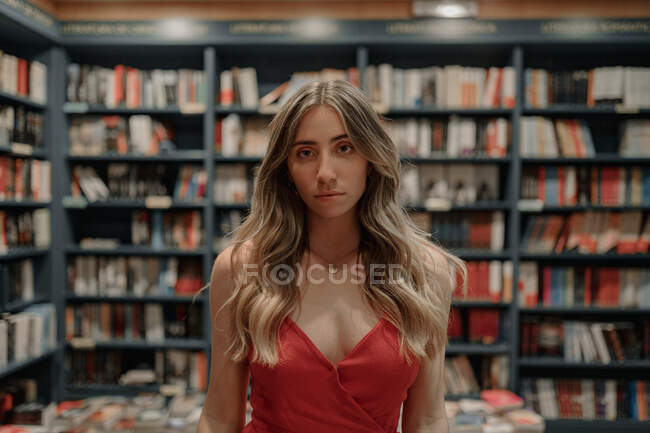 Elegante joven sin emociones en vestido de fiesta de pie contra la mesa con una variedad de libros de texto en la librería - foto de stock