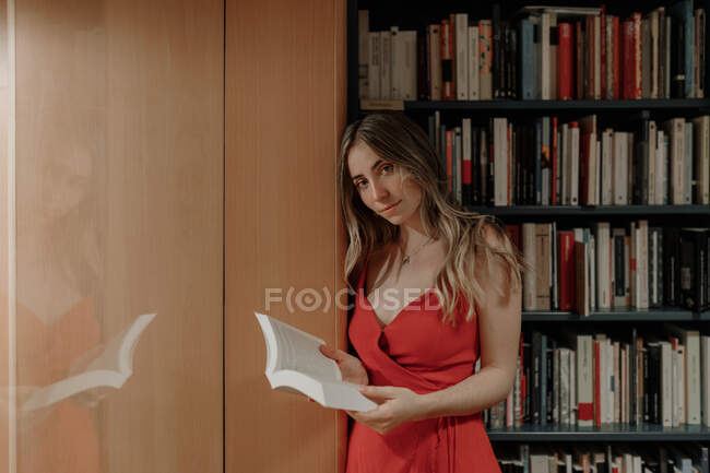 Vista lateral da jovem fêmea em sundress vermelho com livro aberto em pé na livraria e olhando para a câmera — Fotografia de Stock