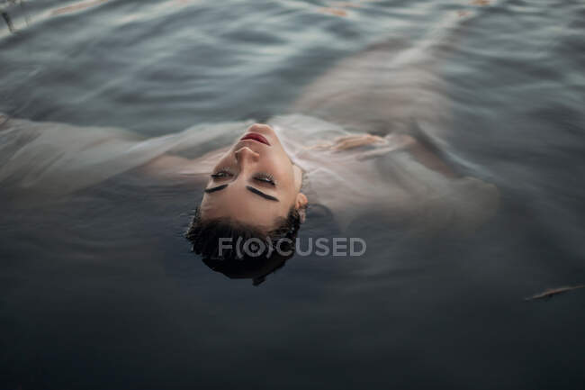 From above serene young brunette in wet panties and translucent white cloth lying with eyes closed on tranquil dark pond water — Stock Photo