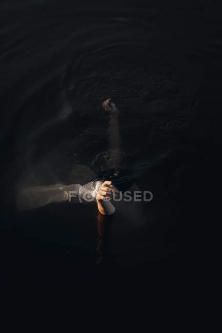 From above side view of unrecognizable female traveler covering face with textile while standing with raised arms in water at dusk — Stock Photo