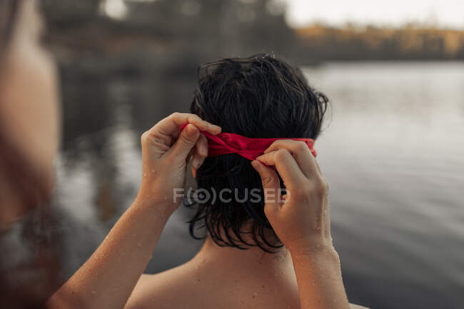 Crop anonymous female tourist tying red blindfold on head of partner against rippled water during trip — Stock Photo