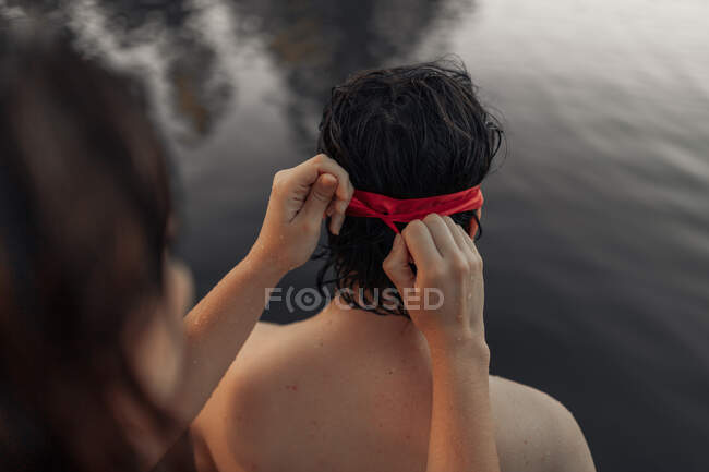 Ritaglia anonima turista femmina legando benda rossa sulla testa del partner contro l'acqua increspata durante il viaggio — Foto stock