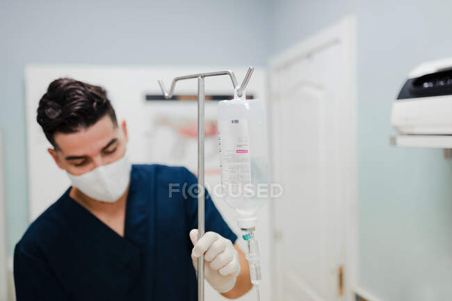 Anonymous young male nurse in respiratory mask installing drop counter with solution in transparent bag in hospital — Stock Photo