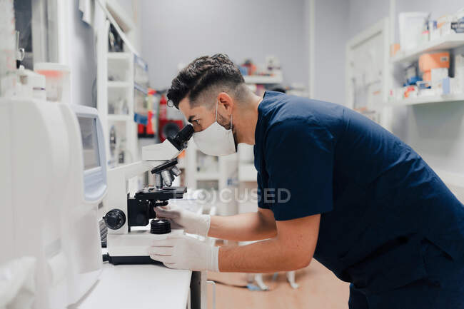 Вид сбоку анонимного мужчины-медика в форме и маске с помощью микроскопа во время работы в лаборатории — стоковое фото