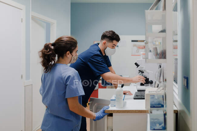 Side view of crop anonymous male medic in uniform and mask using microscope while working in lab near woman colleague — Stock Photo