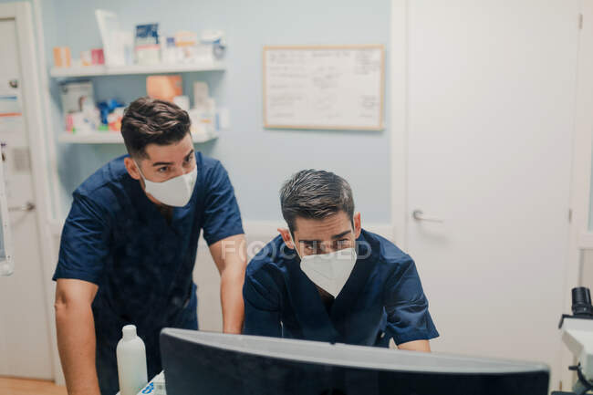 Анонімні молоді лікарі в дихальних масках і уніформах працюють на настільних комп'ютерах у клініці. — стокове фото
